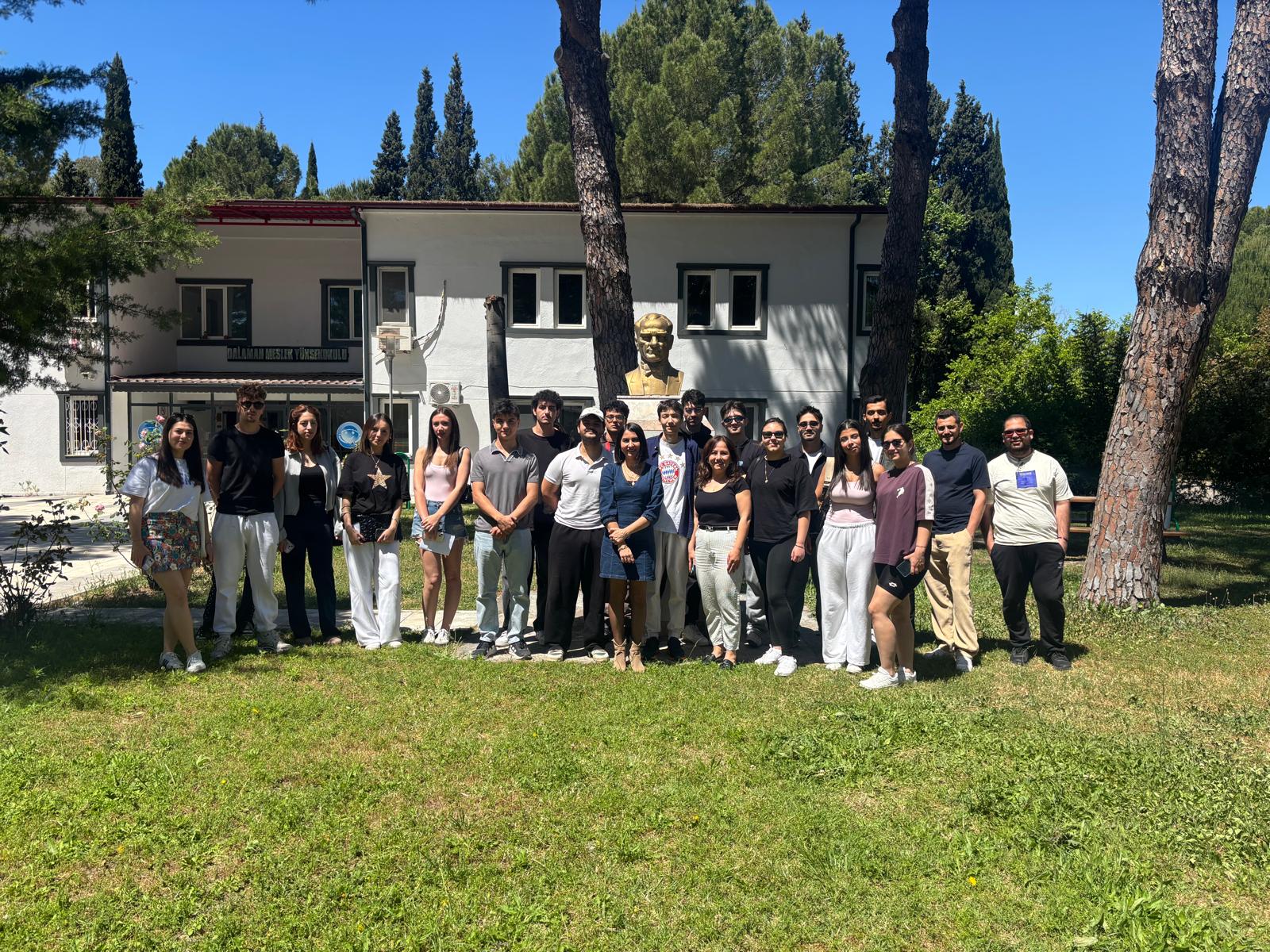 In the photo, taken on a grassy area, a large group is standing side by side. In the background is a white, two-story school building. A bust of Mustafa Kemal Atatürk stands in front of the  building. Tall pine trees and greenery can be seen in the surrounding area. The sky is clear and sunny. The group is posing in a line for the camera.